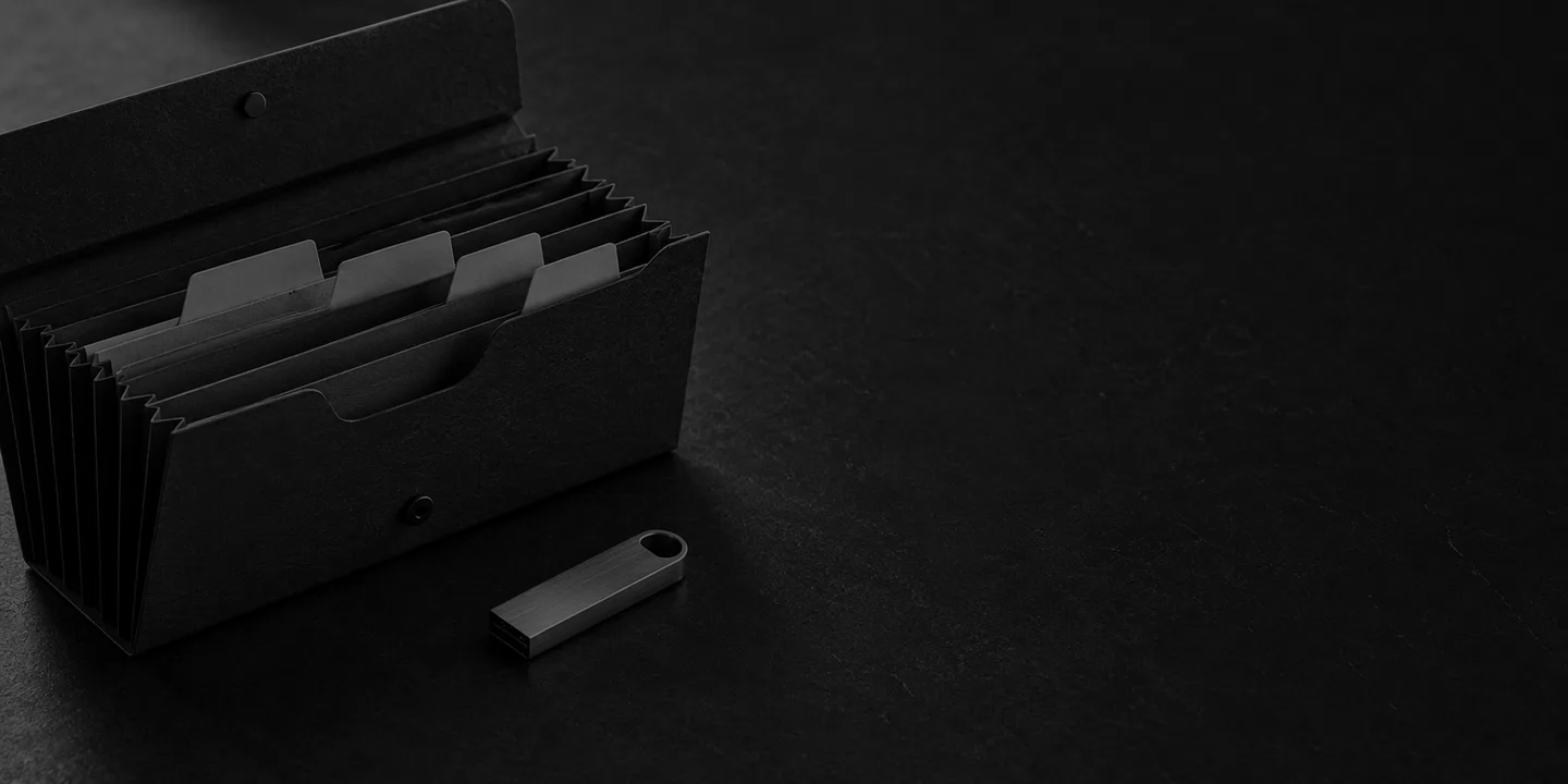 Greyscale conceptual accordion document wallet with blank divider tabs beside an unbranded metal USB drive on a dark desk.
