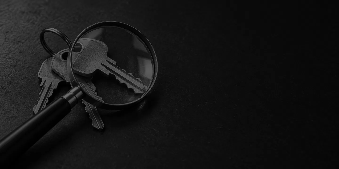Greyscale conceptual magnifying glass resting over ordinary metal keys on a dark desk.